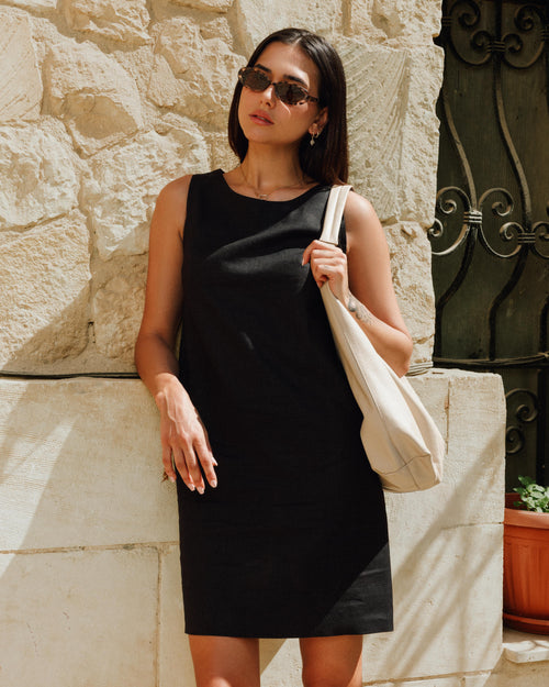 Woman in a black dress holding a beige bag against a stone wall.